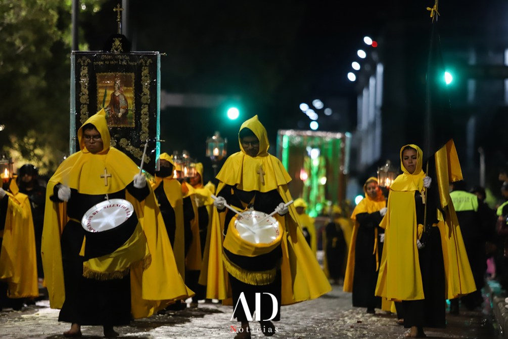 Participantes en una procesión nocturna, vestidos con capas amarillas y capuchas, sosteniendo un estandarte y tocando tambores en una calle iluminada.