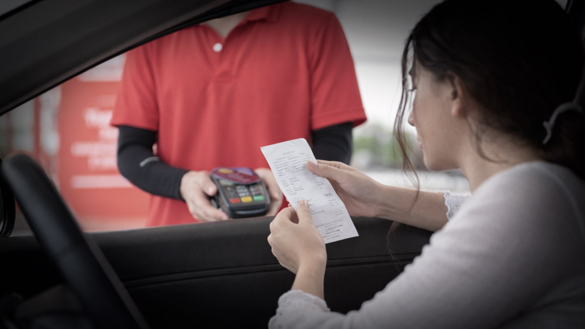 Una mujer recibe un recibo de un empleado que está en el exterior del automóvil. Ambos parecen estar en un servicio de autoservicio.
