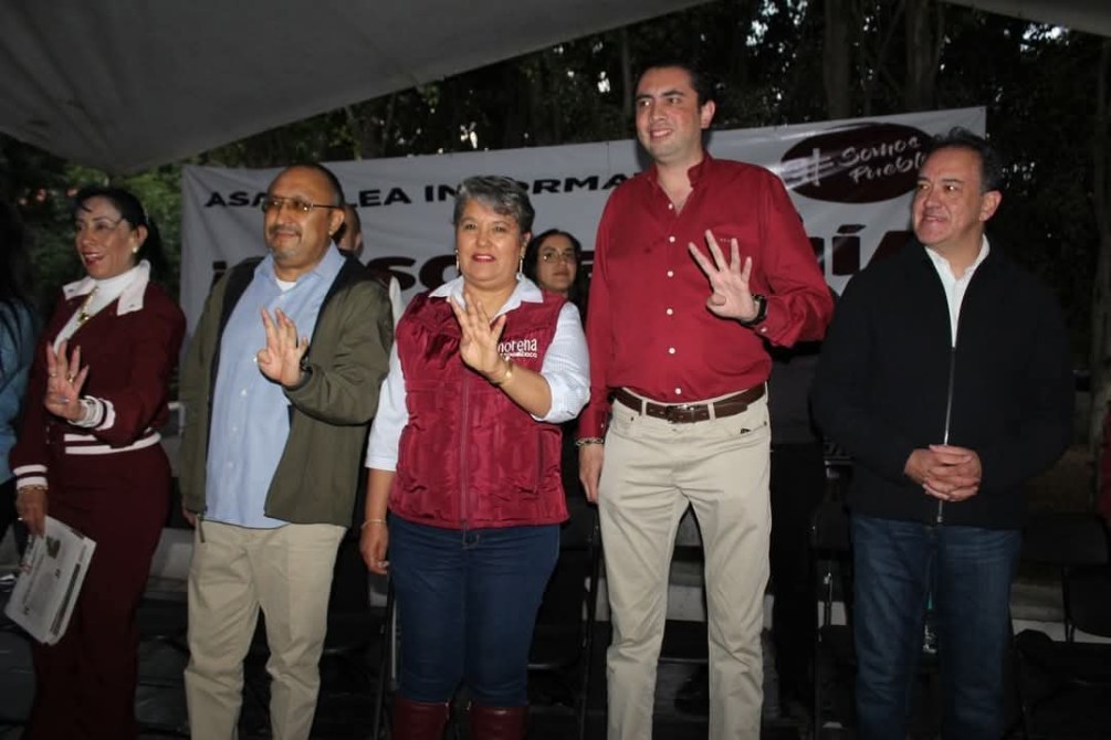 Grupo de personas posando con gestos de triunfo en un evento comunitario. Algunos visten chaquetas y prendas de varios colores, mientras se encuentra un banner de fondo.