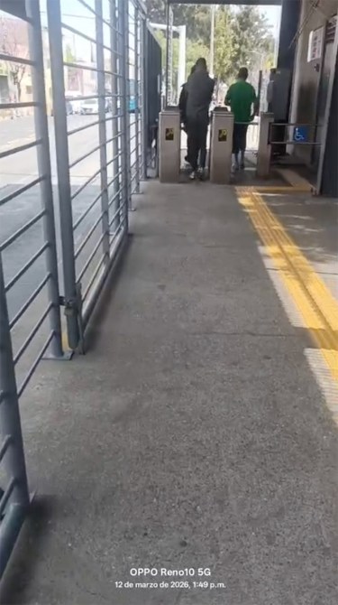 Dos personas caminando hacia una estación de transporte público, pasando por torniquetes en un área urbana.