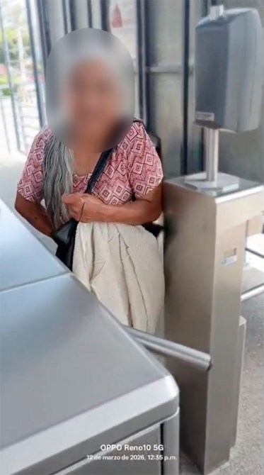 Persona mayor con cabello largo y canoso, sosteniendo una bolsa, en un acceso de transporte público.