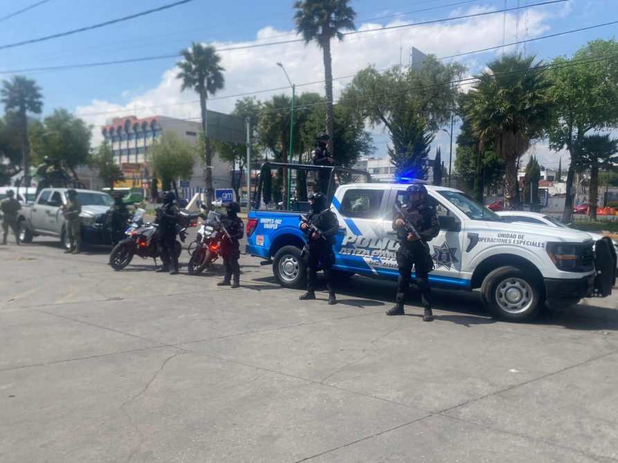Grupo de policías armados en una calle, con vehículos policiales y motos. Un vehículo policial azul y blanco está estacionado mientras algunos agentes se encuentran de pie y otros en la parte trasera.