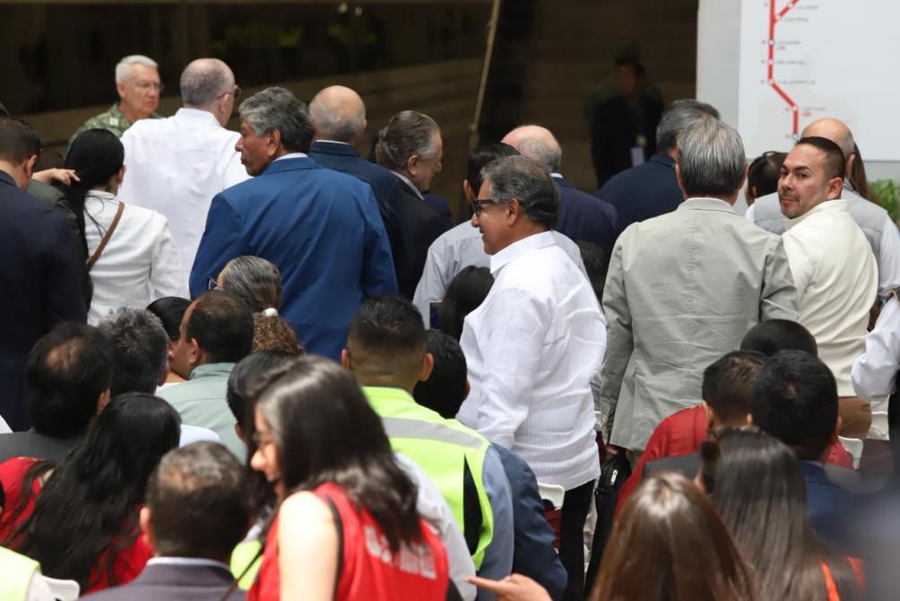 Grupo de personas de pie en un evento, muchas de espaldas, con diferentes atuendos formales y chaquetas. Algunos llevan chalecos reflectantes, mientras que otros parecen pertenecer a una ceremonia o presentación.