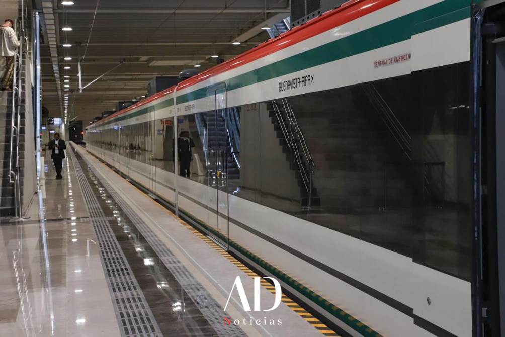 Estación de tren moderna con un tren de color blanco y verde en la plataforma, personas caminando y escaleras al fondo.