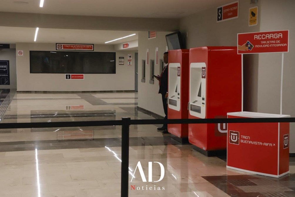 Interior de una estación de tren con máquinas expendedoras rojas y un quiosco de recarga. Hay señalización en las paredes y se observa una persona de pie al fondo.