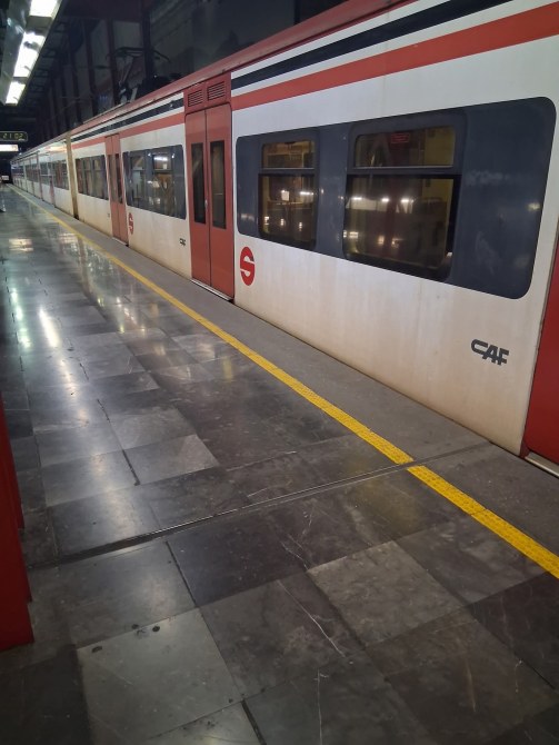 Un tren de cercanías estacionado en una plataforma con un suelo de mármol. La parte de la estación es moderna con iluminación artificial.
