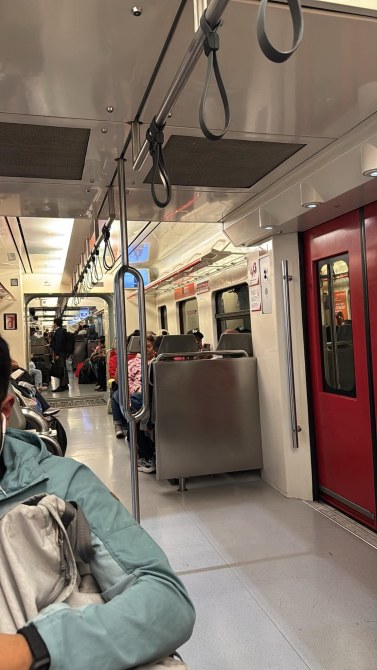 Interior de un tren con asientos ocupados y algunas personas de pie. Hay agarraderas en el techo y una puerta roja al fondo.