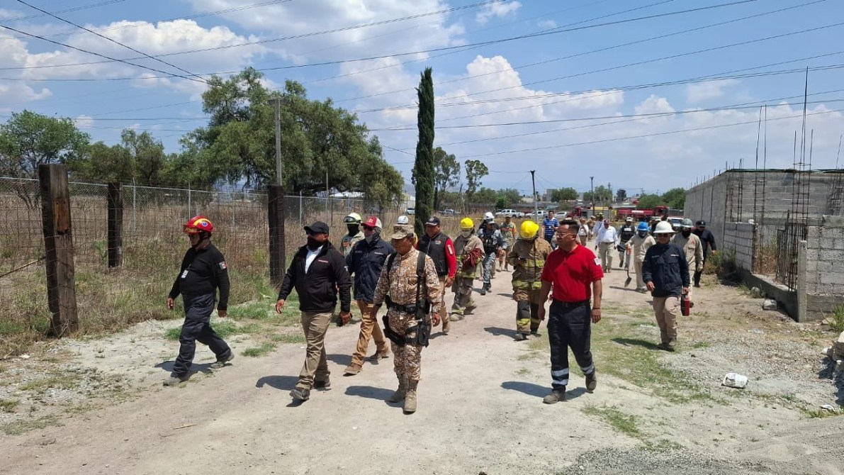 Grupo de personas caminando por un camino de tierra, algunas con cascos y equipo de seguridad, en un entorno rural con árboles y cercas.