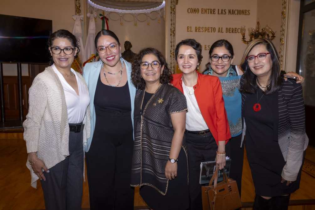 Seis mujeres posando juntas en un evento, sonriendo y vistiéndose de manera formal, con fondo decorativo que incluye la bandera de México.