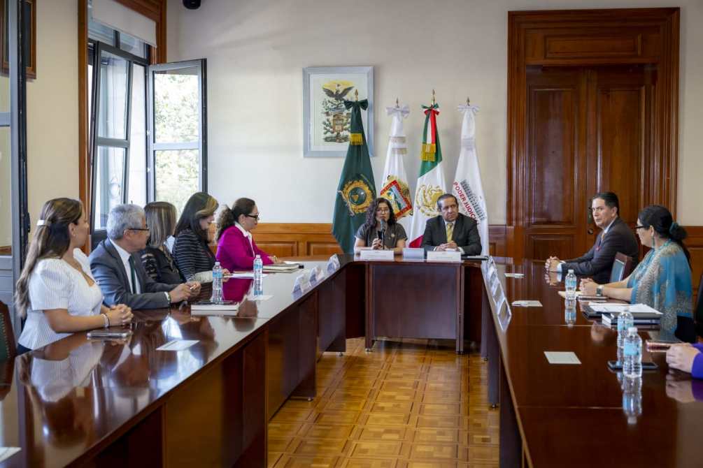 Reunión en una sala de conferencias con funcionarios sentados alrededor de una mesa, algunos sosteniendo botellas de agua. En la pared hay banderas y un cuadro enmarcado.