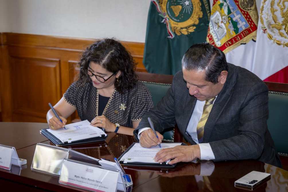 Dos personas firmando documentos en una mesa, con banderas visibles de fondo.