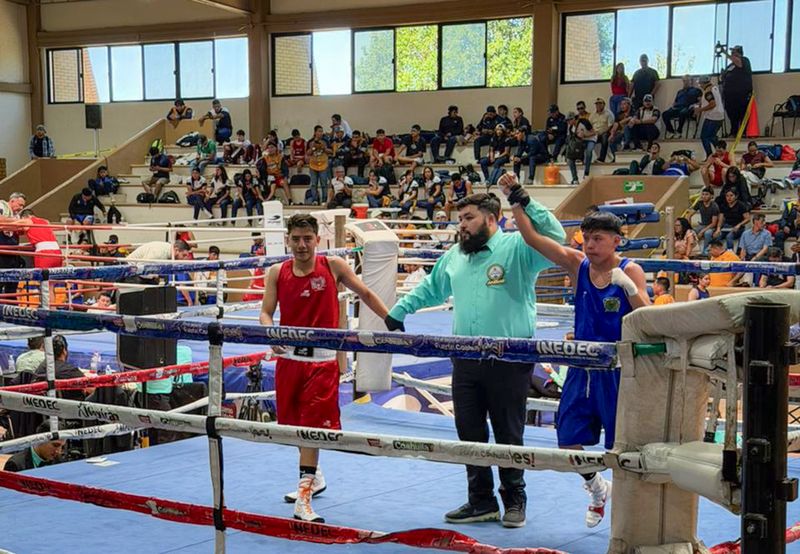 Un árbitro levanta la mano de un boxeador con camiseta roja mientras el otro boxeador en camiseta azul espera en el ring, con un público observando en las gradas.