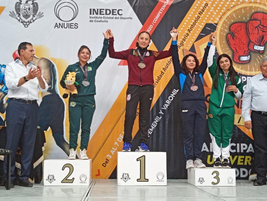 Cuatro mujeres en un podio de medallas durante una competencia de boxeo, levantando las manos en señal de victoria. La atleta en el centro lleva una medalla de oro y sostiene la mano de sus compañeras, que tienen medallas de plata y bronce. Al fondo, un banner con motivos de boxeo y el logotipo de la competencia.