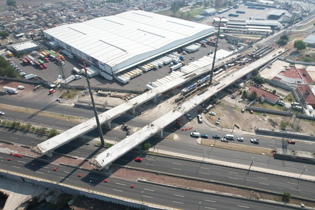 Vista aérea de una construcción de puente elevado sobre una carretera, con maquinaria y trabajadores en el sitio, y un gran edificio industrial al fondo.