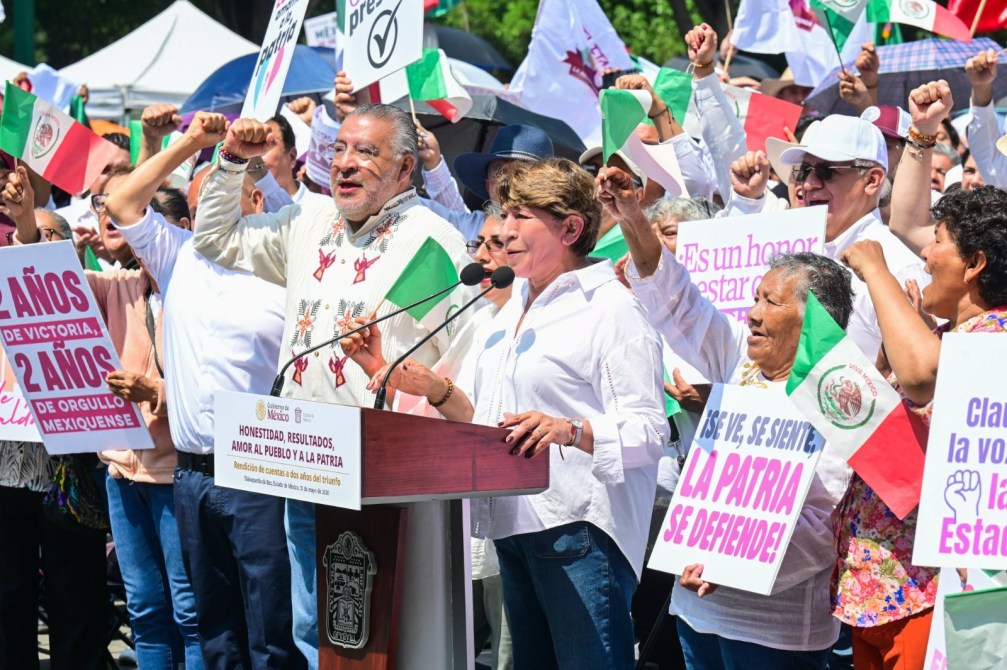 Manifestación con participantes levantando pancartas y banderas mexicanas, en un evento que celebra dos años de gobierno, con oradores en un podio.