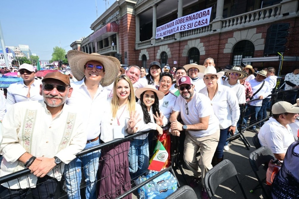 Grupo de personas sonrientes en un evento al aire libre, detrás de una valla, vistiendo camisas blancas y sombreros, con cartel de bienvenida a la gobernadora en el fondo.