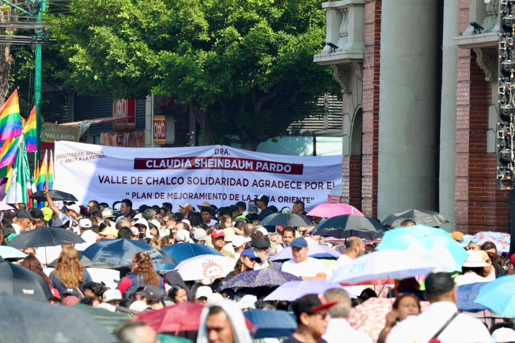 Multitud de personas en una marcha sosteniendo un cartel que dice 'Dra. Claudia Sheinbaum Pardo, Valle de Chalco Solidaridad agradece por el plan de mejoramiento de la zona oriente'. Se observan paraguas de diferentes colores y banderas multicolores.