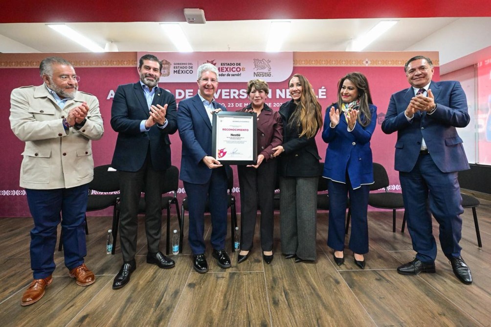 Un grupo de seis personas, incluyendo a un hombre sosteniendo un reconocimiento de Nestlé, posan para la foto en un evento de entrega de reconocimientos. Todos aplauden y están vestidos de manera formal, con un fondo decorativo que incluye el logotipo de Nestlé.