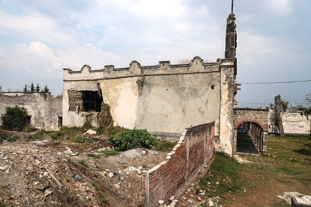 Edificio en ruinas con paredes agrietadas y enrejadas, rodeado de maleza y escombros.