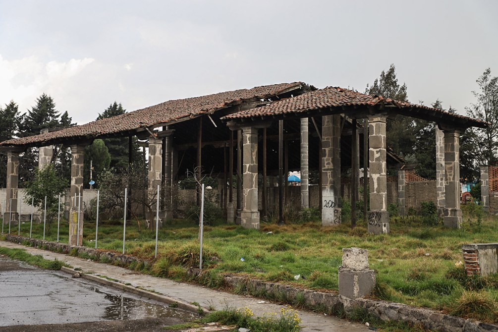 Edificio en ruinas con techo de tejas, columnas expuestas y vegetación crecida alrededor.