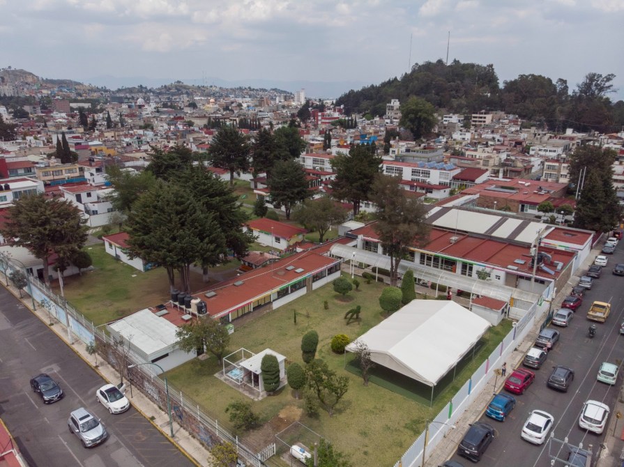 Vista aérea de un área urbana con edificios y casas de techos rojos, incluyendo un parque con árboles y una carpa blanca en el centro.
