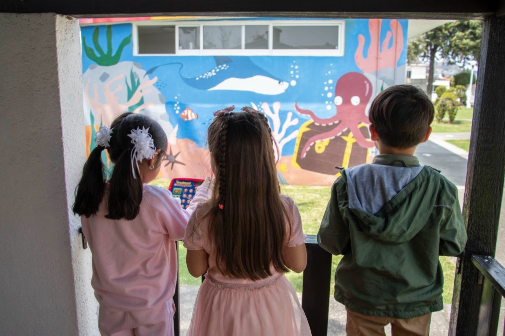 Tres niños observan un mural colorido de un pulpo y vida marina desde una entrada.