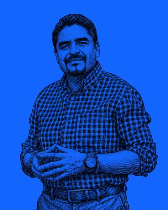 Retrato de un hombre sonriente de pie, vestido con una camisa de cuadros, sobre un fondo azul.