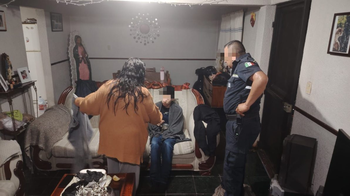 Una mujer de pie interactuando con un niño sentado en un sofá, mientras un oficial de policía observa en la sala de una casa decorada con imágenes y muebles sencillos.