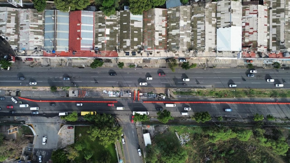 Vista aérea de una carretera con vehículos en movimiento, flanqueada por edificios de techos metálicos y árboles.