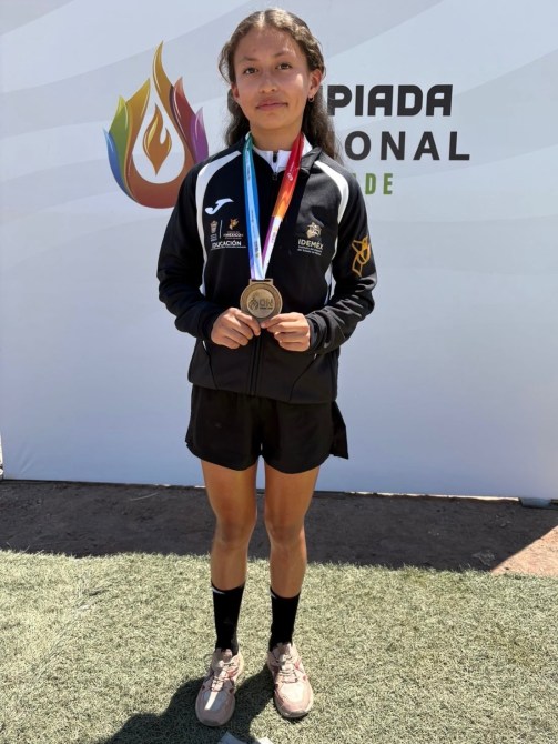 Niña sosteniendo una medalla de oro en un evento deportivo, vestida con un uniforme deportivo negro, de pie sobre un césped artificial, con un fondo que muestra el logotipo de la Olimpiada Nacional.