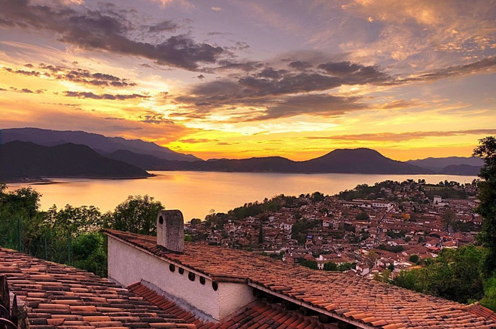 A scenic sunset view over a lake and mountainous landscape, with a town featuring terracotta rooftops in the foreground.