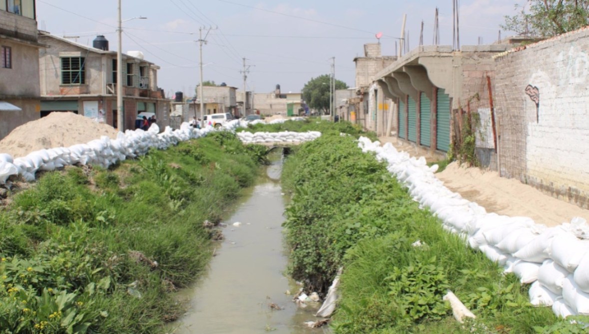 Vista de un canal con agua contaminada rodeado de vegetación y sacos de arena, ubicado en un área urbana.