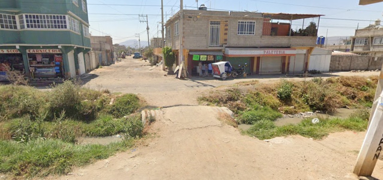 Vista de una calle en un área urbana, con tiendas y casas a ambos lados. Se observa un camino de tierra y vegetación, además de un pequeño puente sobre un canal.