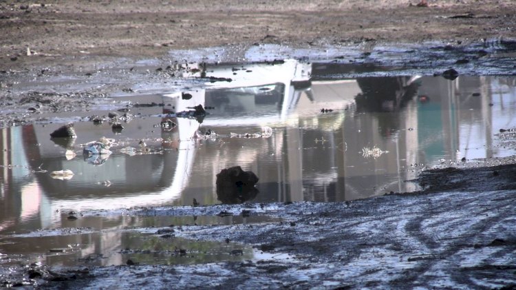 Charco de agua en un terreno sucio con escombros y reflejo de un vehículo.
