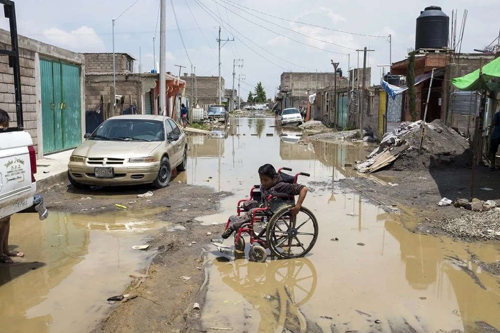 Niño en silla de ruedas navegando por una calle inundada en un vecindario urbano.