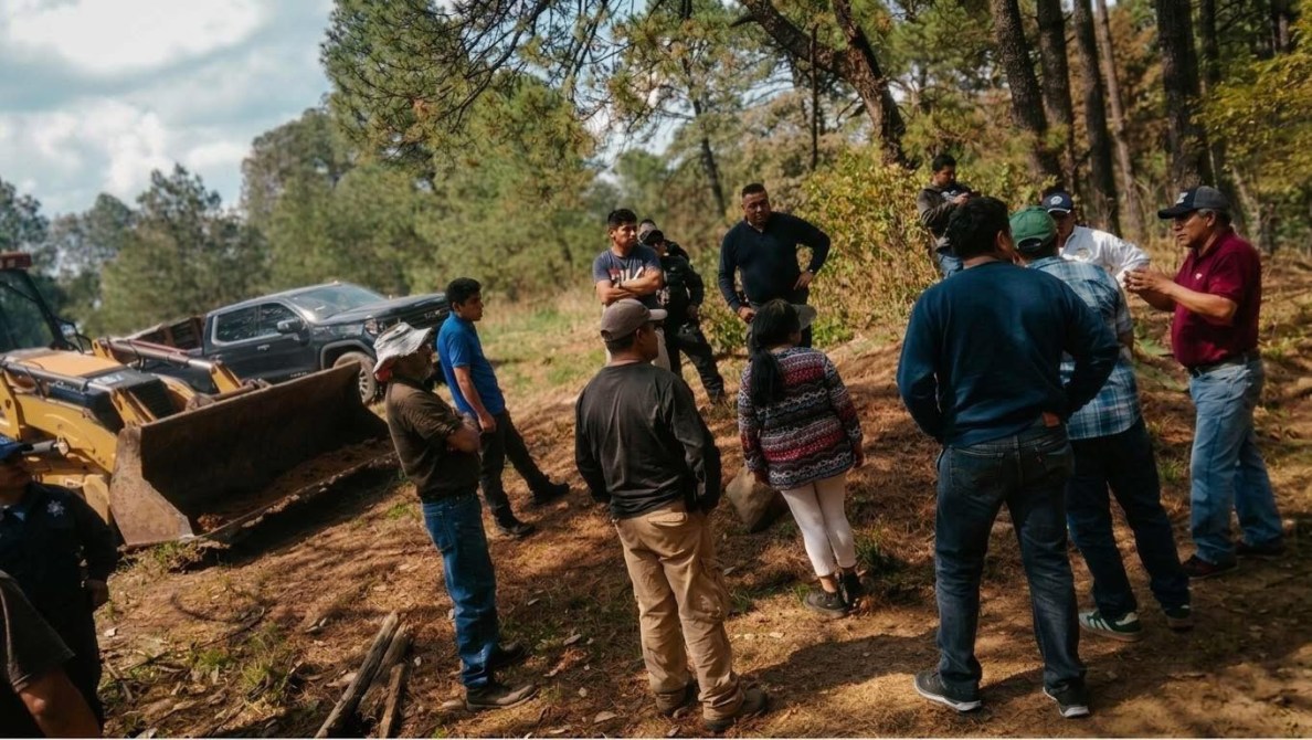 Grupo de personas conversando en un área boscosa, con maquinaria de construcción y vehículos en el fondo.