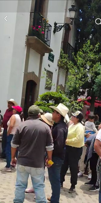 Grupo de personas en una plaza al aire libre, algunas usando sombreros, con un edificio blanco y vegetación de fondo.