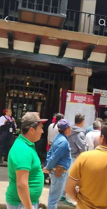 Una multitud de personas en fila frente a un edificio, con un cartel informativo visible en la entrada.
