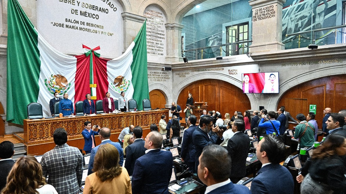 A legislative assembly session in Mexico, featuring officials standing at a podium beneath a large Mexican flag backdrop, with attendees seated and standing in the foreground.