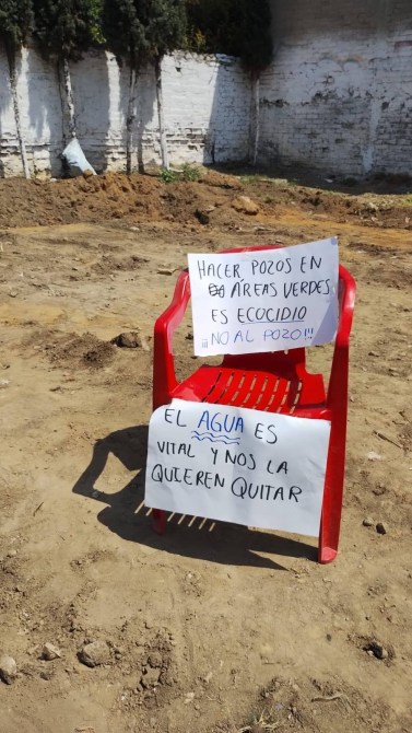 A red plastic chair with two signs on it sitting on bare soil. The first sign reads, 'Making wells in green areas is ecocide, NO to the well!' in Spanish. The second sign states, 'Water is vital and they want to take it from us.'