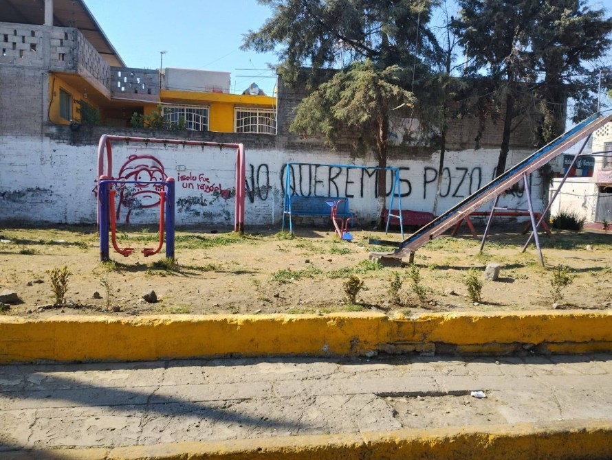 A playground scene featuring two swings and a slide, with a grassy area and some shrubs. A wall in the background displays graffiti that reads 'No queremos pozo'.