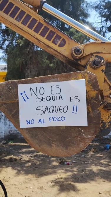 A construction vehicle arm with a sign that reads '¡NO ES SEQUIA ES SAQUEO!! NO AL POZO' advocating against extraction.