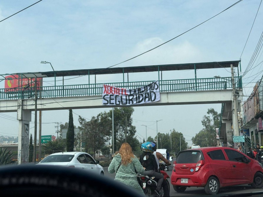 A banner reading 'Xochimilco y la Seguridad' hangs from a pedestrian overpass above a busy street, with vehicles and motorcyclists visible below.