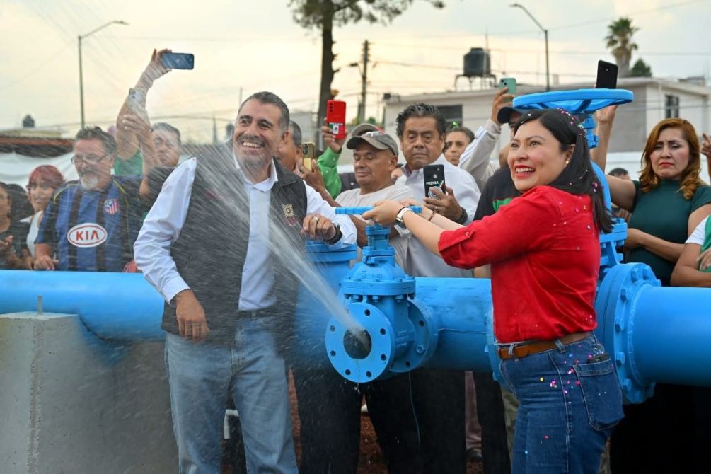 A joyful event where two individuals are joyfully inaugurating a large blue water valve, spraying water. A crowd of onlookers is gathered, capturing the moment with their smartphones.