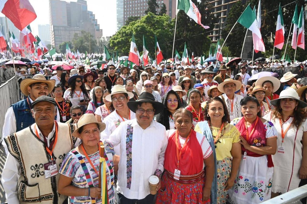 Grupo de personas en un evento al aire libre, vistiéndose con trajes tradicionales, con banderas de México ondeando al fondo.