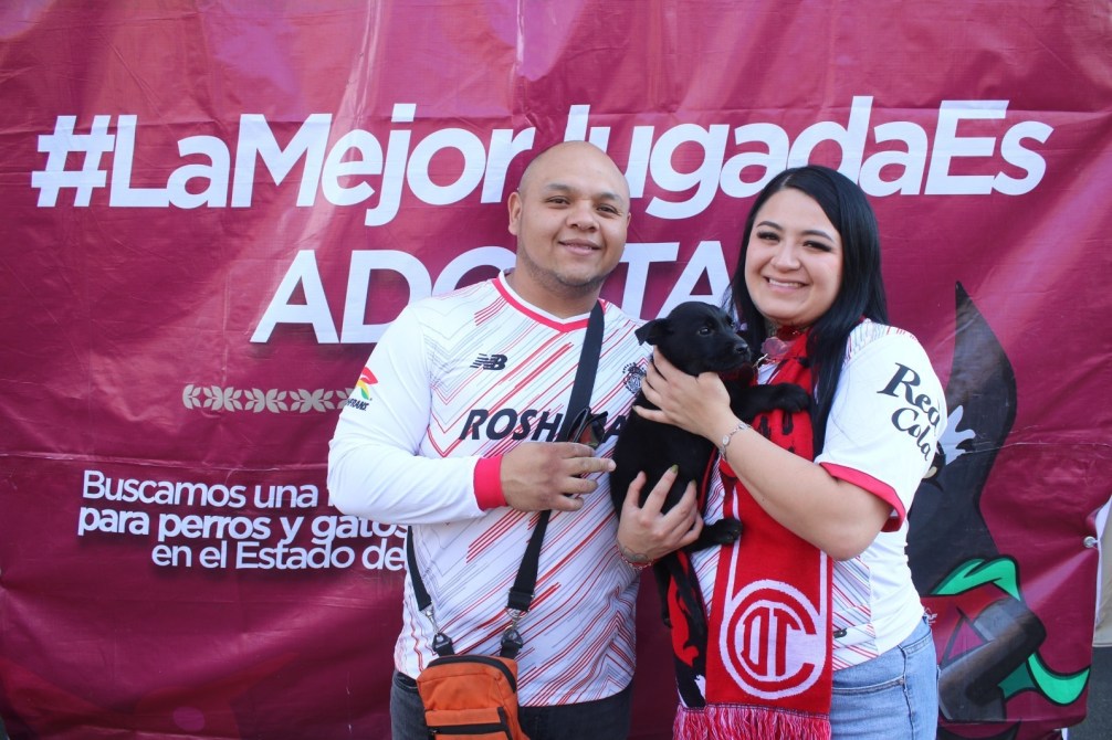 Una pareja sonriente sostiene un perro pequeño frente a un fondo con el hashtag #LaMejorJugadasEs y un mensaje sobre la adopción de mascotas.