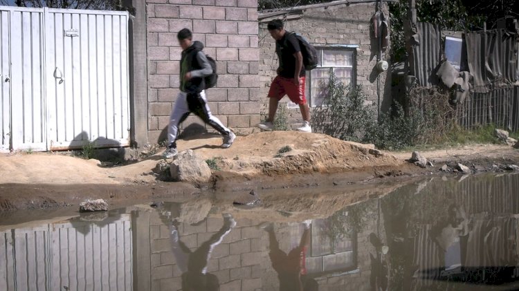 Dos adolescentes caminan por un sendero junto a una reflexión en un charco, con una pared de ladrillo y una puerta blanca al fondo.