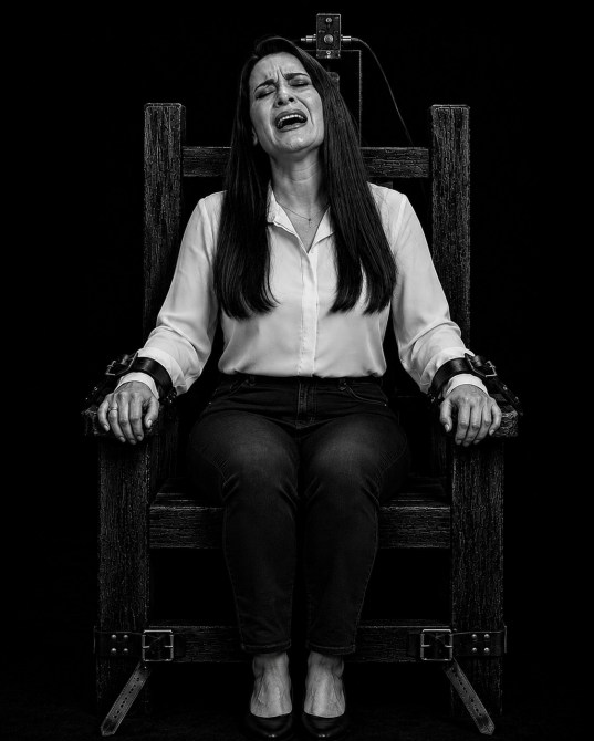 Una mujer con pelo largo y oscuro, vestida con una camisa blanca y pantalones oscuros, sentada en una silla eléctrica, con una expresión de angustia en su rostro, en un fondo negro.