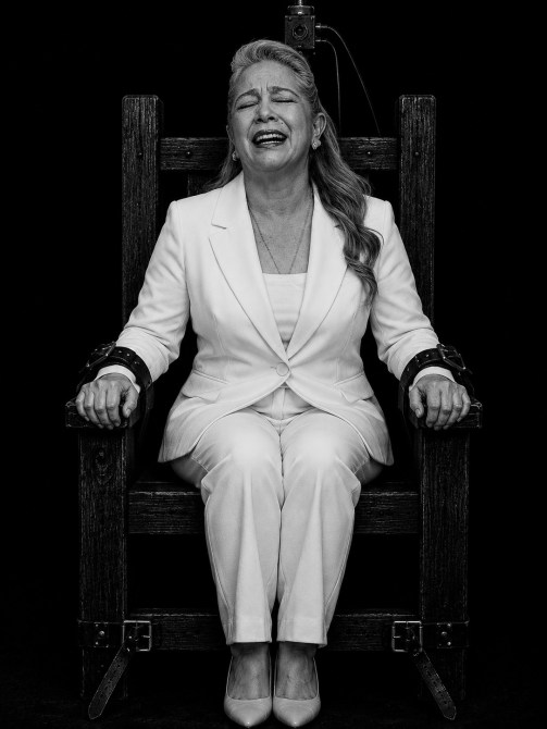 Mujer con cabello largo y suelto, vestida de blanco, riendo mientras está sentada en una silla con grilletes, en un fondo negro.