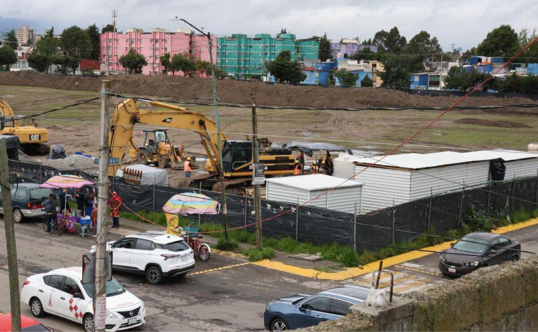 Construcción en un terreno con maquinarias pesadas, incluyendo excavadoras y obreros trabajando, junto a vehículos y puestos de venta en la calle.